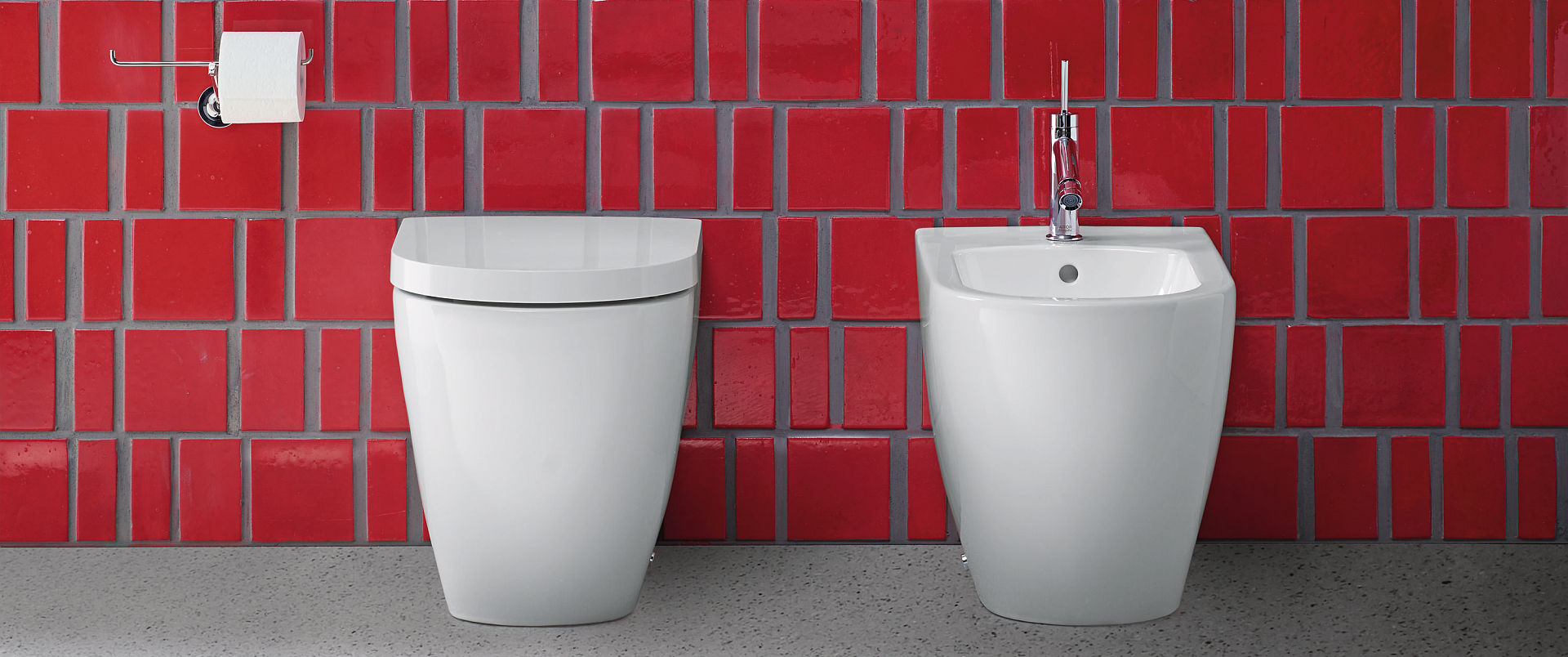 Ein elegantes weißes WC und Bidet, minimalistisch gestaltet, vor einer Wand aus roten Kacheln in kräftigem Glanz.