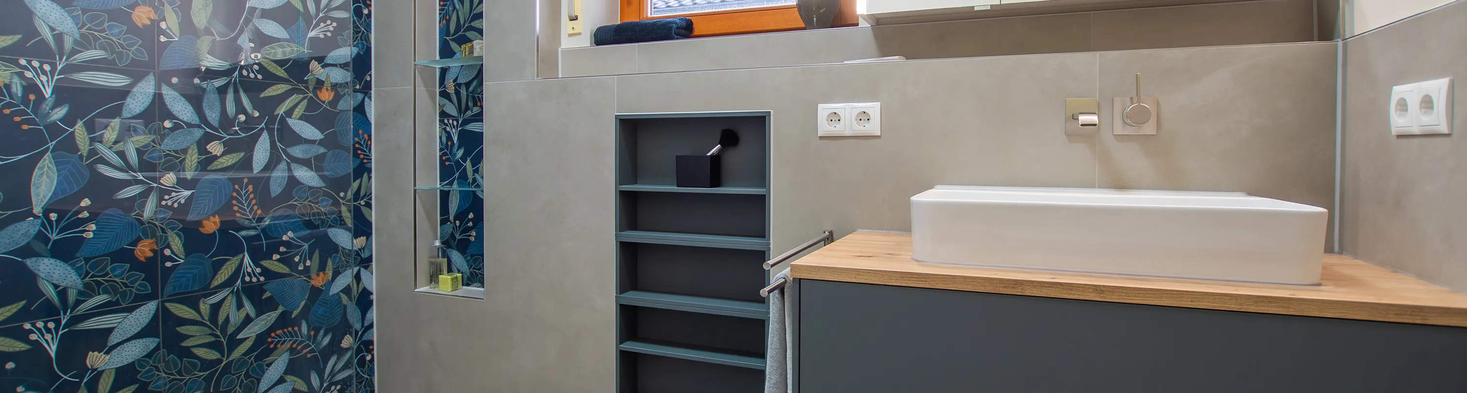 Badezimmer mit großem Duschbereich mit floraler Fliese, sowie einem Waschtisch mit dunkelgrauem Unterschrank. Header Format.