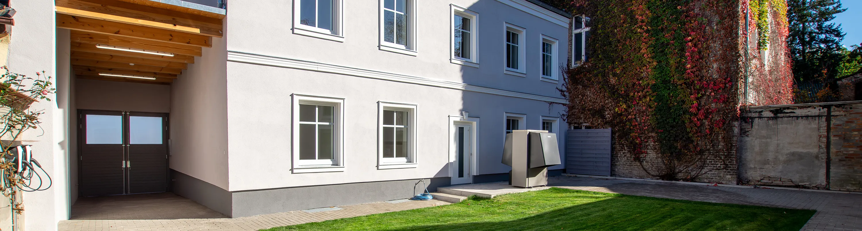 Zweifamilienhaus mit hellgrauer Fassade und Wärmepumpe im Garten.