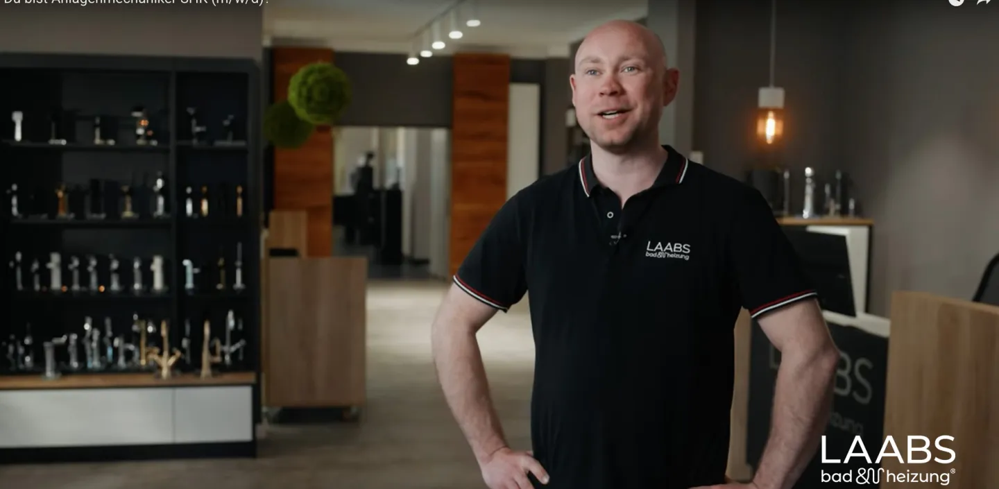 Ein Mann in einem modernen Bäder-Showroom steht selbstbewusst mit verschränkten Armen und trägt ein schwarzes Poloshirt mit Firmenlogo. Im Hintergrund sind hochwertige Armaturen und Sanitärprodukte in Regalen ausgestellt, die den professionellen Charakter des Raums unterstreichen. Warme Beleuchtung und Holzelemente sorgen für eine einladende Atmosphäre. Das Bild vermittelt Kompetenz und Handwerksqualität im SHK-Bereich.