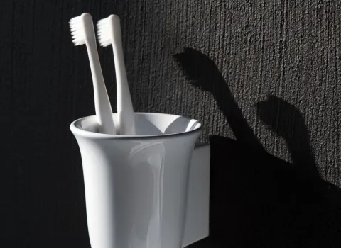 Zwei weiße Zahnbürsten stehen in einem weißen, glänzenden Becher, der an einer dunkelgrauen, strukturierten Wand befestigt ist. 