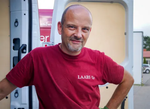 Ein Badmonteur SHK der Firma LAABS in rotem Shirt Firmen-Logo lehnt an der offenen Tür seines Transporters.