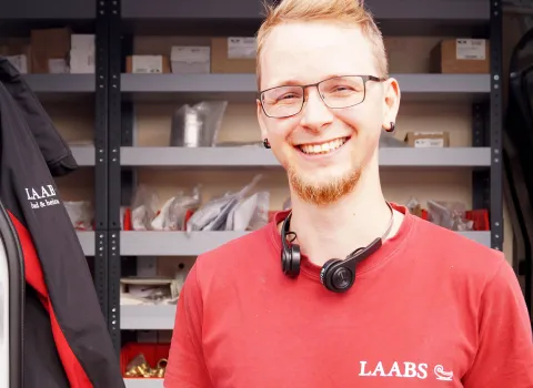Ein junger Kundendienstmonteur SHK der Firma Laabs mit Brille und Kopfhörern um den Hals lächelt in die Kamera.