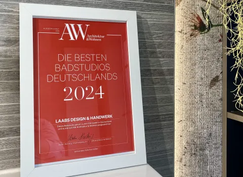 Eine gerahmte Auszeichnung von AW Architektur & Wohnen für Laabs Design & Handwerk als eines der besten Badstudios Deutschlands 2024 steht dekorativ vor einer modernen Holzstrukturwand.