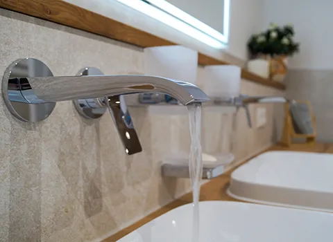 Waschtisch, Detailaufnahme: Eine Wandarmatur in chrom von Hansgrohe, aus der Wasser läuft.