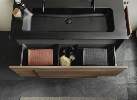 Ein Waschtisch in Holzoptik mit offenem Auszug präsentiert ein klares Ordnungssystem mit Fächern für Handtücher und Pflegeprodukte.