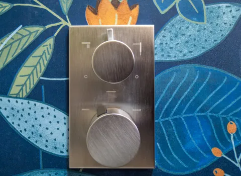 Nahaufnahme eines Edelstahl-Duschthermostats von DORNBRACHT vor einer blau-floralen Duschwand.
