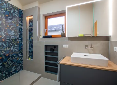 Modernes Badezimmer mit floral gestalteter Duschwand, großem beleuchtetem Spiegel, offenem Regalfach und breitem Waschtisch auf Holzplatte.