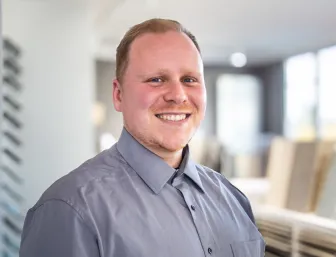 Junger Mann lächelt in die Kamera, er hat ein graues Hemd an und steht in einer Fliesenausstellung.