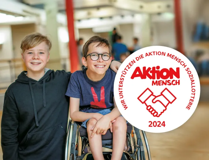 Zwei lachende Jungen, einer im Rollstuhl, umarmen sich freundschaftlich, während ein Logo der Aktion Mensch Soziallotterie 2024 die Unterstützung für Inklusion betont.