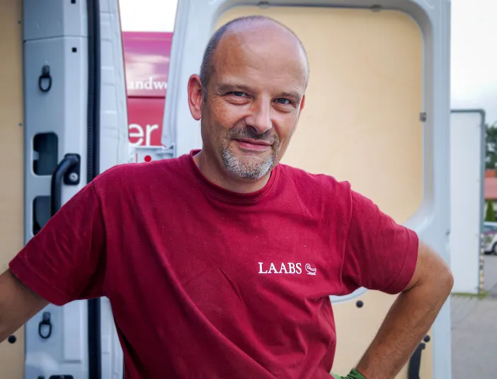 Ein Badmonteur SHK der Firma LAABS in rotem Shirt Firmen-Logo lehnt an der offenen Tür seines Transporters.